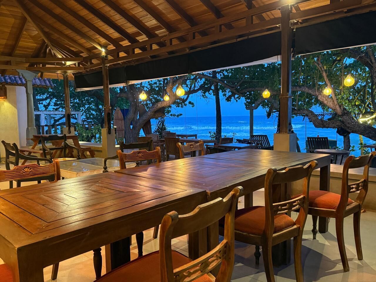 Un área de comedor al aire libre con mesas y sillas de madera, con vista a un cuerpo de agua.