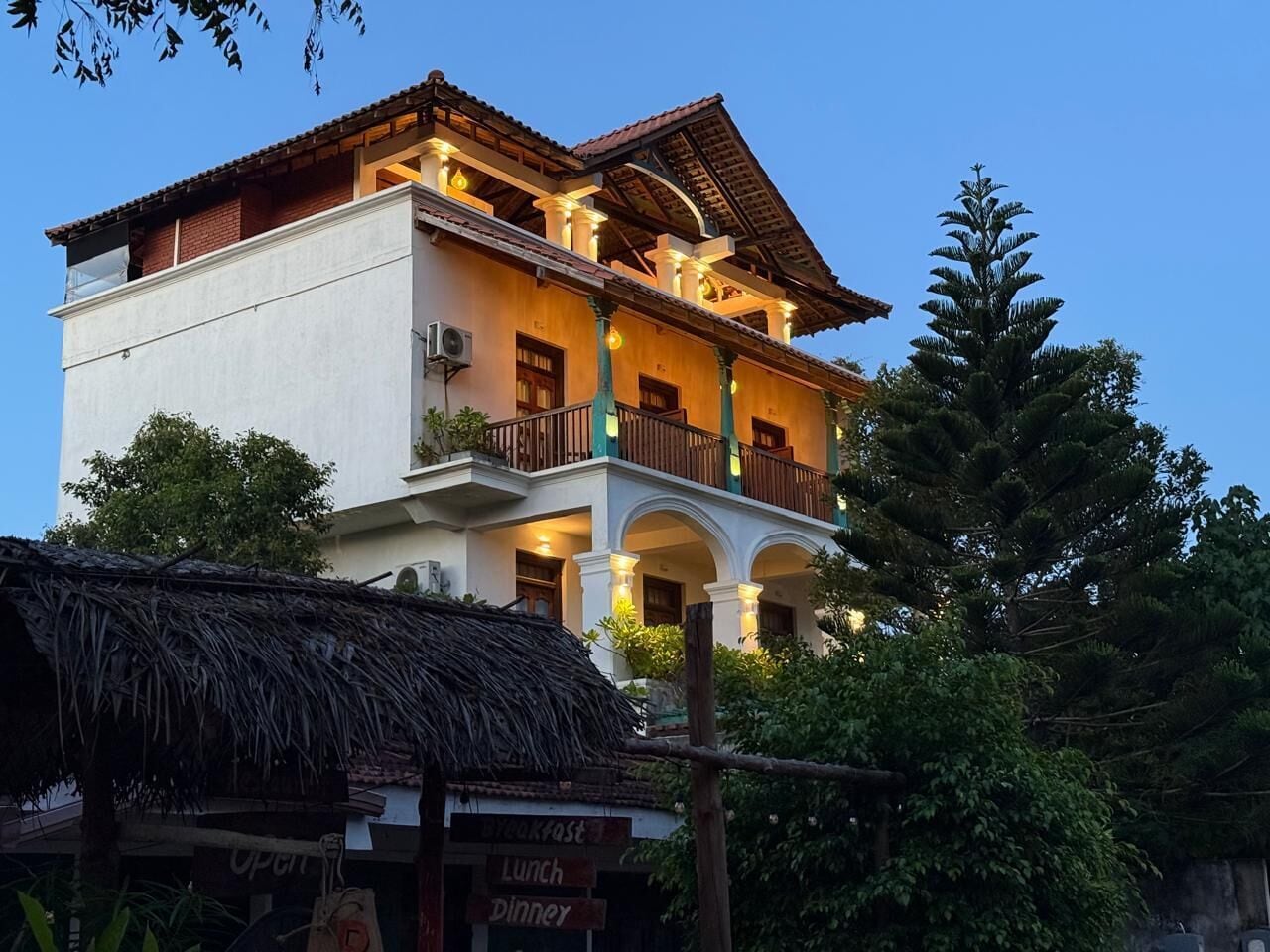 Un edificio de varios pisos con balcones, una estructura de paja en primer plano y un cartel que indica servicios de restauración.