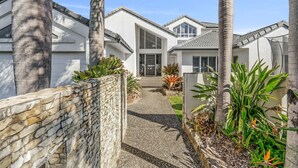 A modern house with a stone wall, palm trees, and a pathway leading to the entrance. - Waterfront Oasis (Minyama)