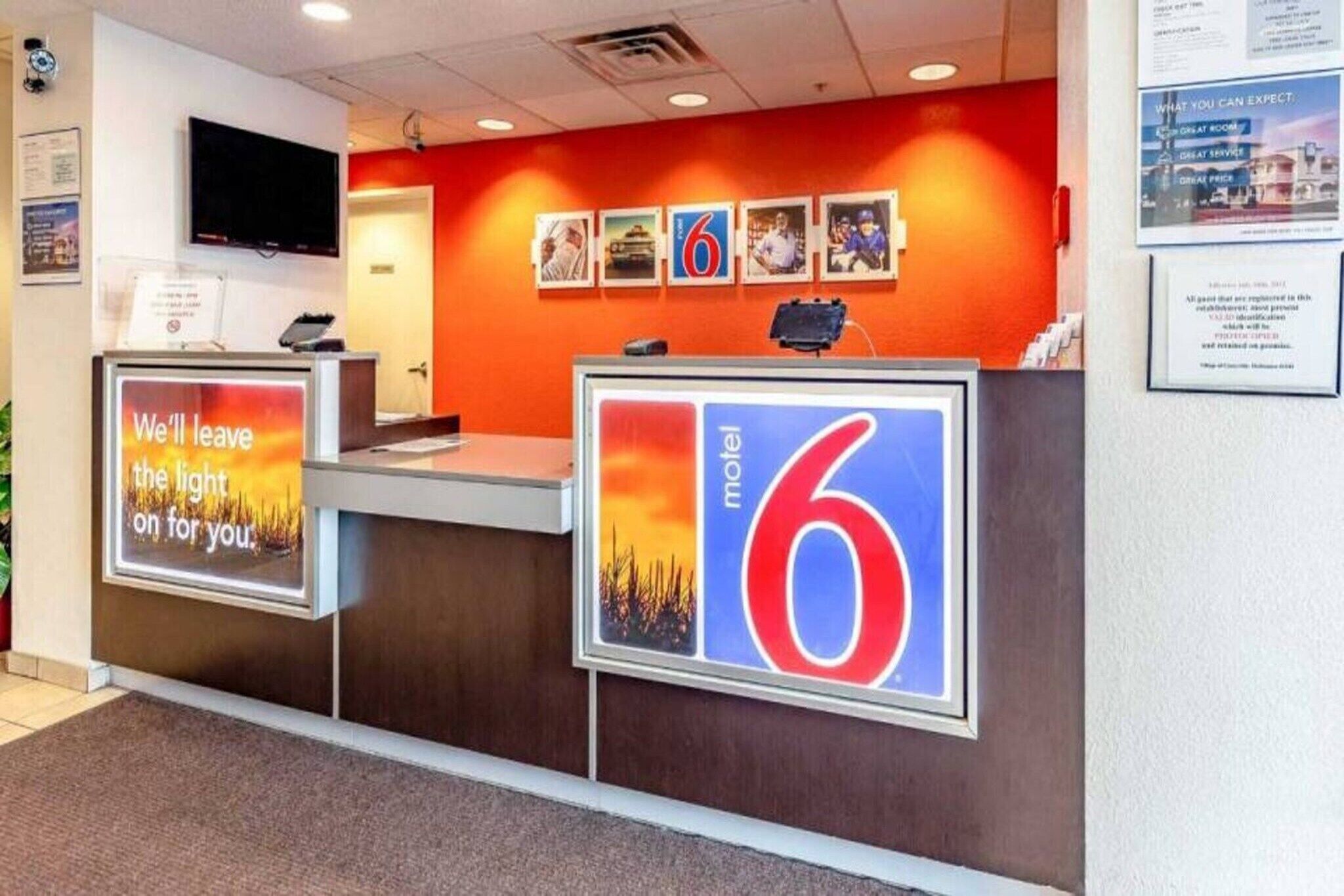 A hotel reception desk with a large number '6' sign, a TV mounted on the wall, and a framed picture of a sunset.