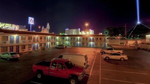 Un hotel de varios pisos, un estacionamiento con varios autos y un edificio destacado con un letrero iluminado al fondo.