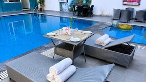 Zona junto a la piscina con sillones de mimbre, una mesa lista para comer y una piscina inflable pequeña.