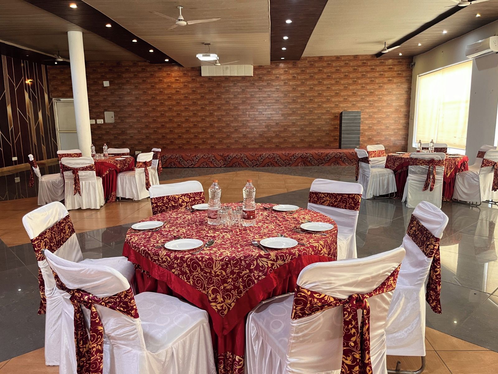 A banquet hall with round tables covered in red tablecloths, white chairs with red sashes, and a brick wall in the background.