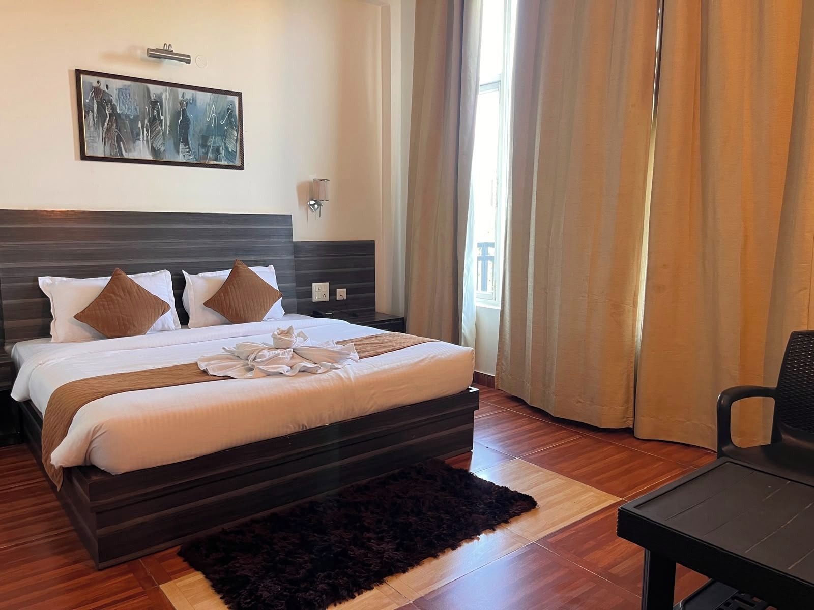 A hotel room with a large bed, a dark headboard, a wall-mounted artwork, a bedside table, a chair, and a window with curtains.