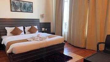 A hotel room with a large bed, a dark headboard, a wall-mounted artwork, a bedside table, a chair, and a window with curtains.