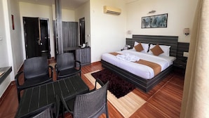 Habitación de hotel con una cama, un escritorio, dos sillas, una alfombra oscura y un cuadro en la pared.