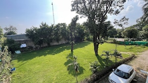 Un espacio al aire libre con césped, donde se ve un coche blanco estacionado en un camino de tierra, rodeado de árboles y un edificio con techo inclinado.