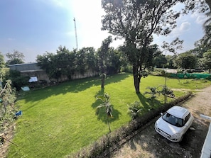 Un espacio al aire libre con césped, donde se ve un coche blanco estacionado en un camino de tierra, rodeado de árboles y un edificio con techo inclinado.