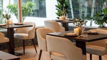 A dining area with tables and chairs, large windows, and indoor plants.