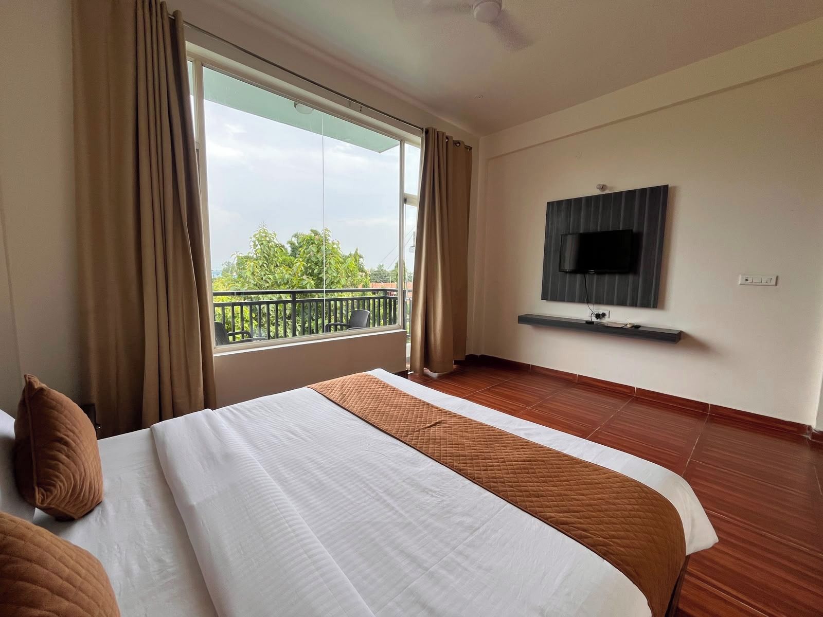 A hotel room with a bed, a TV mounted on the wall, and a balcony with a view of greenery.