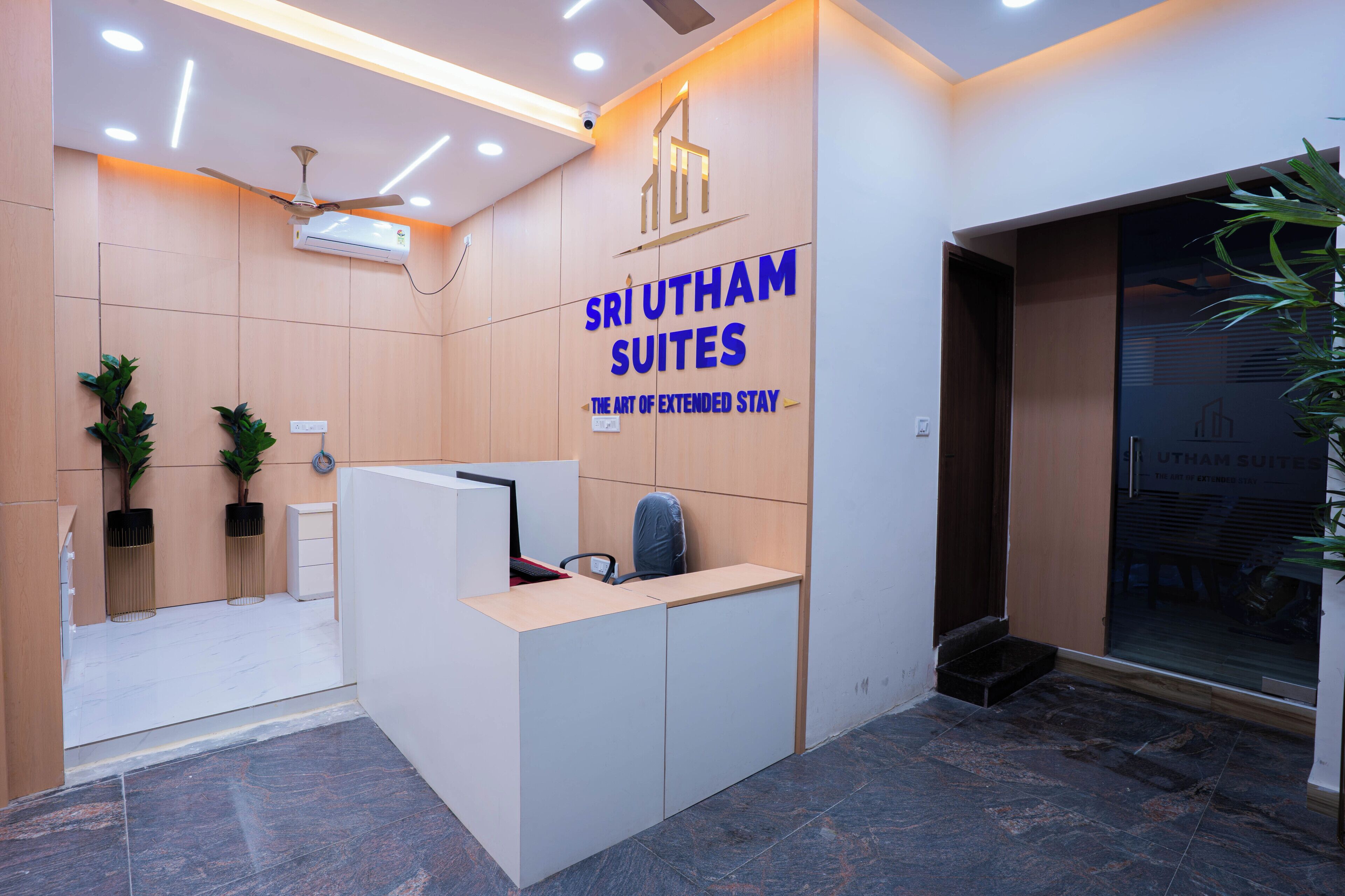 A reception desk with a sign for Sri Utham Suites. There is a ceiling fan, a plant, and a wall-mounted air conditioner.