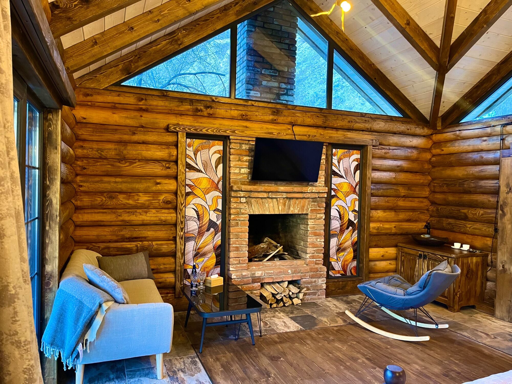 A log cabin interior with a fireplace, a sofa, a rocking chair, and a television mounted on the wall.