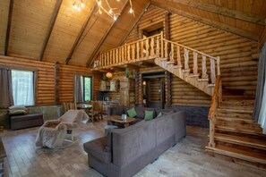 Interior de una cabaña de troncos con una escalera de madera, un sofá y un sillón mecedor.