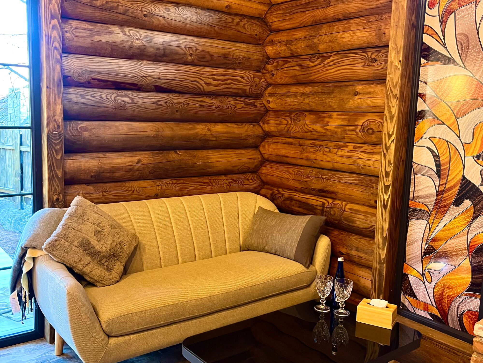 L'intérieur d'une cabane en rondins, avec un canapé, une table en verre sur laquelle se trouvent une bouteille et des verres, et un vitrail.