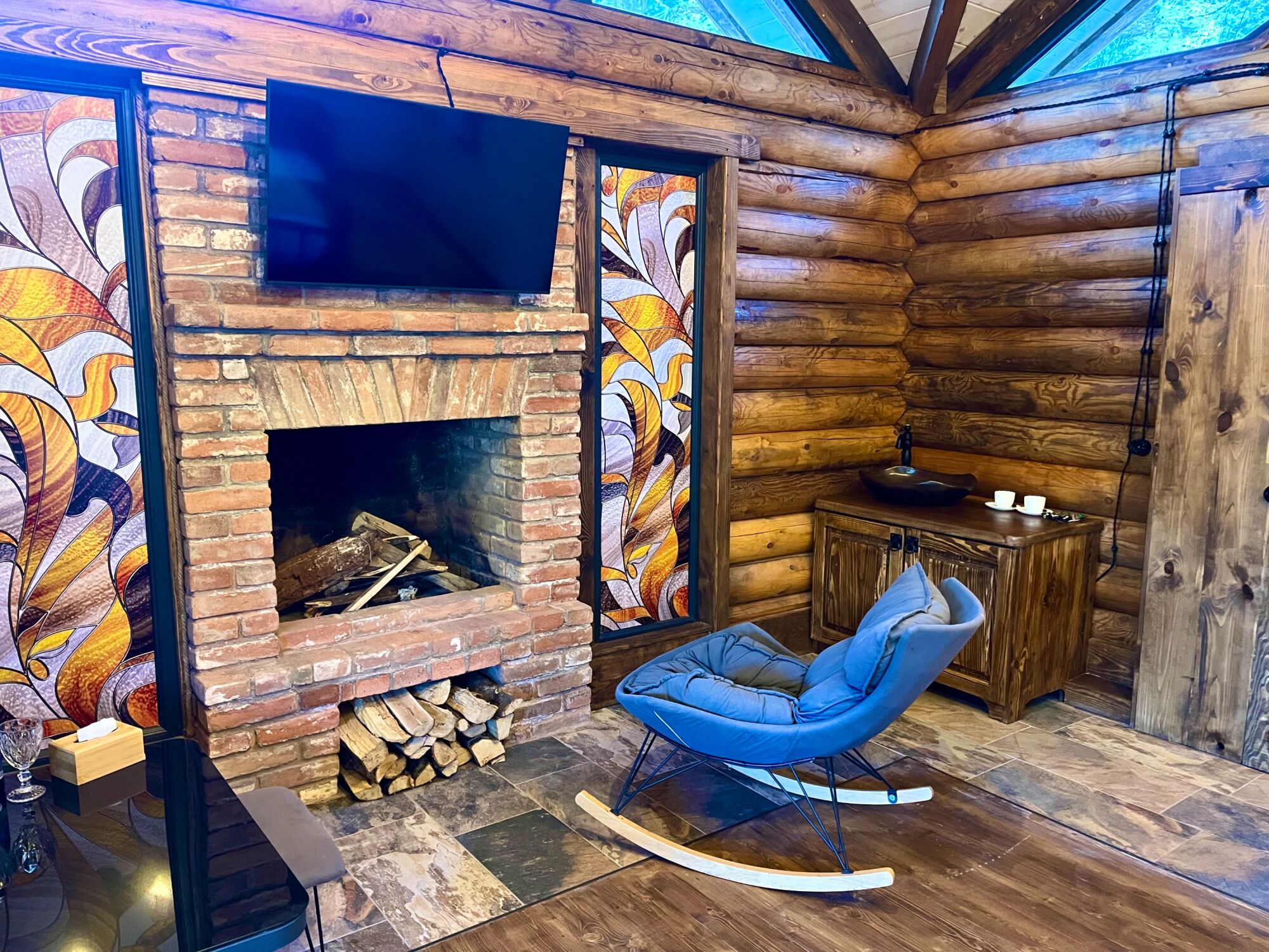 Interior de uma cabana de tronco com lareira, uma cadeira de balanço azul e uma televisão instalada acima da lareira.