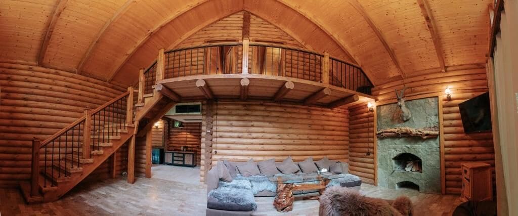 A log cabin interior with a central seating area, a staircase, and a fireplace.