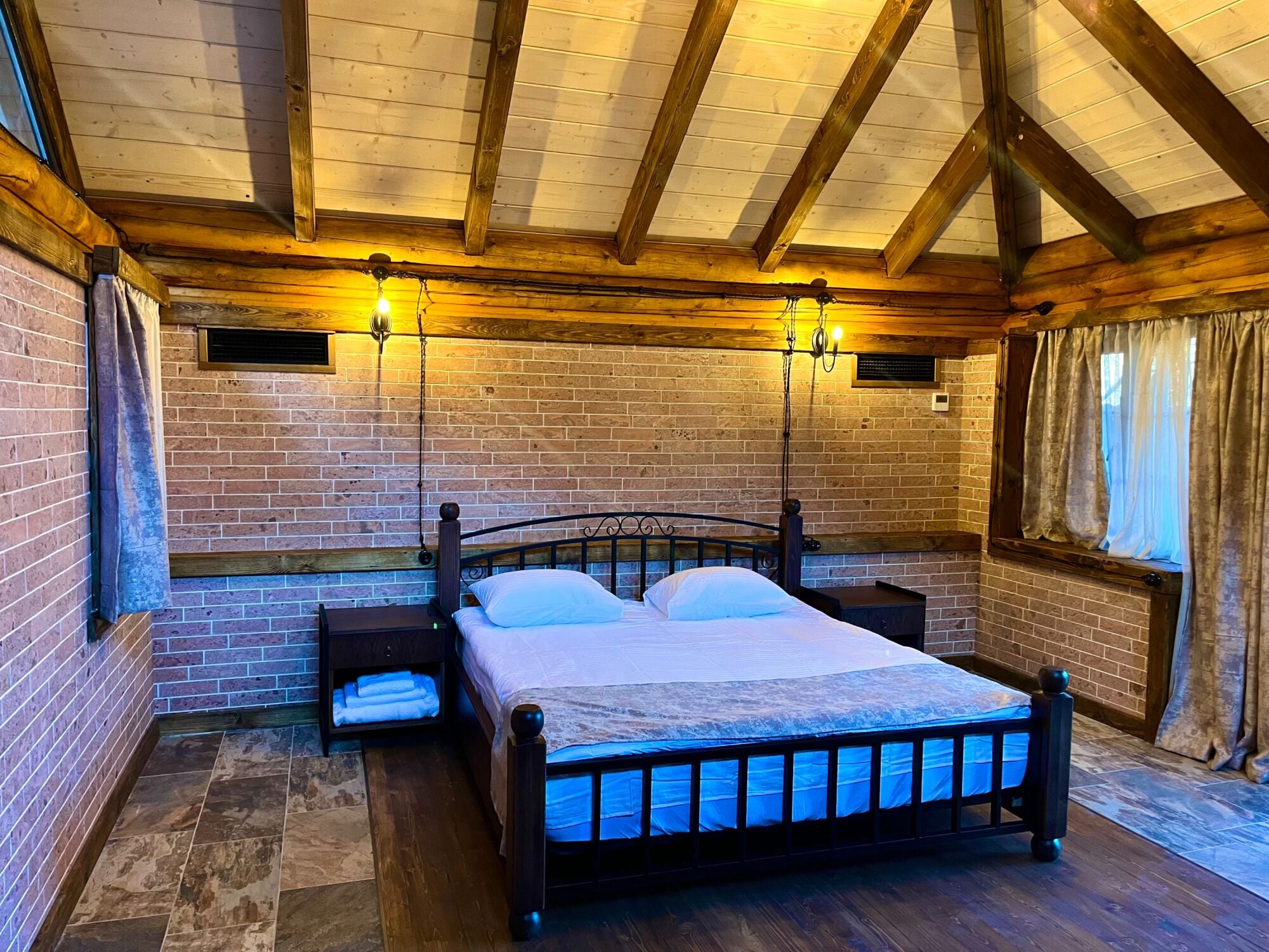 A bedroom with a brick wall, wooden ceiling beams, a bed with a metal frame, two bedside tables, and a window with curtains.