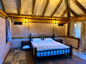 Un dormitorio con pared de ladrillo, vigas de madera en el techo, una cama con estructura metálica, dos mesitas de noche y una ventana con cortinas.