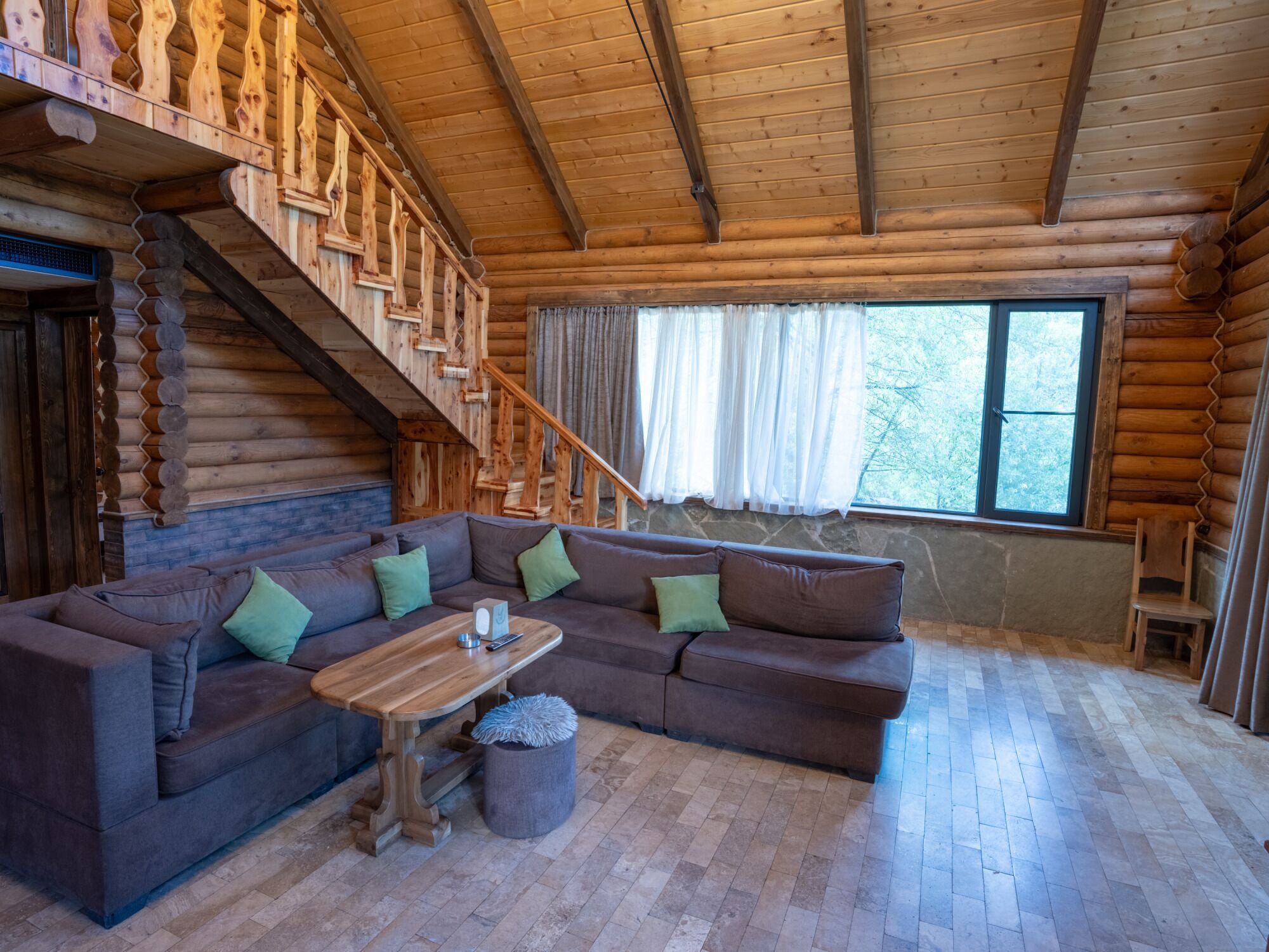 A log cabin interior with a wooden staircase, a sectional sofa, a small table, and a large window.