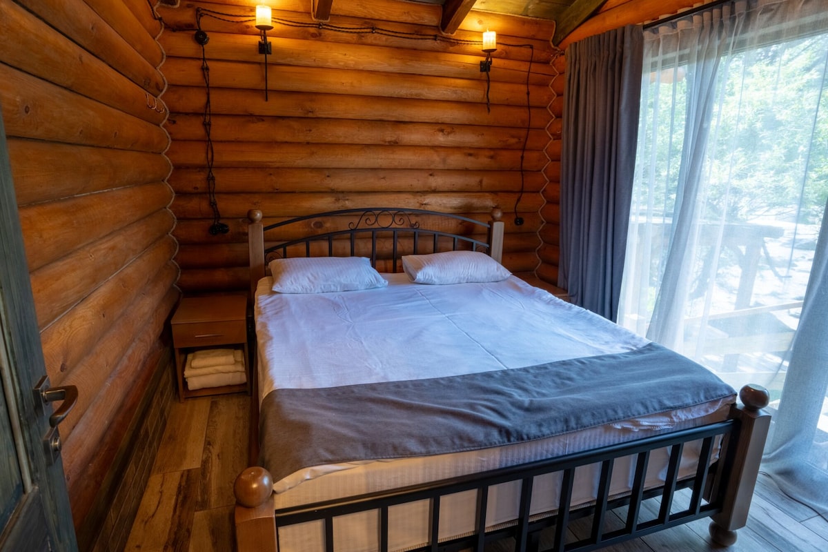 A wooden log cabin room with a bed, bedside lamps, and a window with curtains.