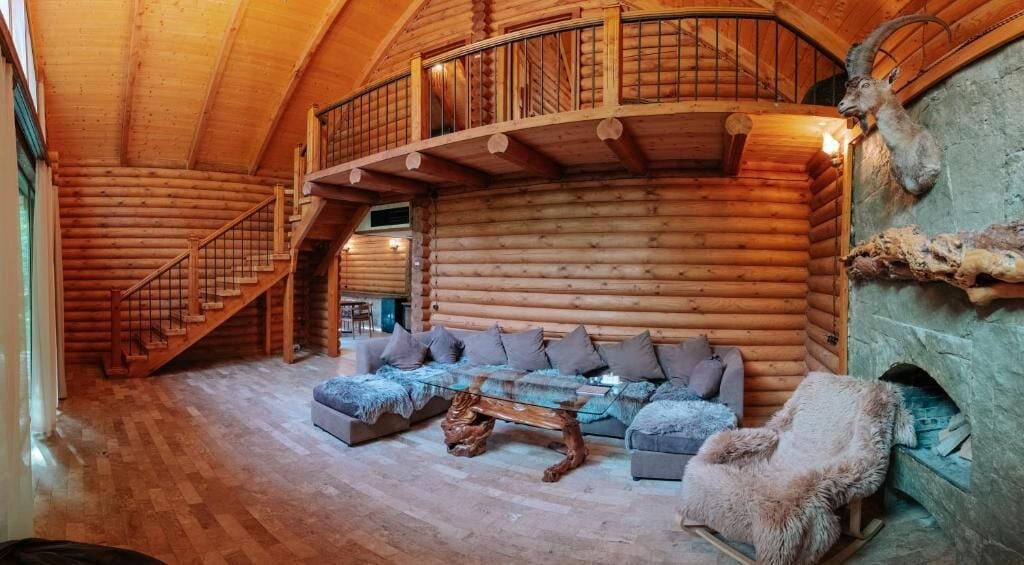 Interior de una cabaña de troncos con escalera, un conjunto de sofás, chimenea y un trofeo de caza en la pared.