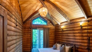 Un dormitorio tipo cabaña de madera con una cama, una ventana con cortinas, una lámpara colgante y una mesita de noche de madera.