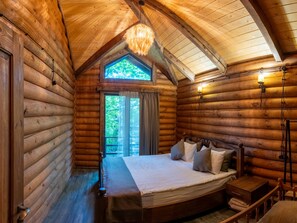 Une chambre dans un chalet en rondins, avec un lit, une fenêtre avec des rideaux, un luminaire suspendu et une table de chevet en bois.