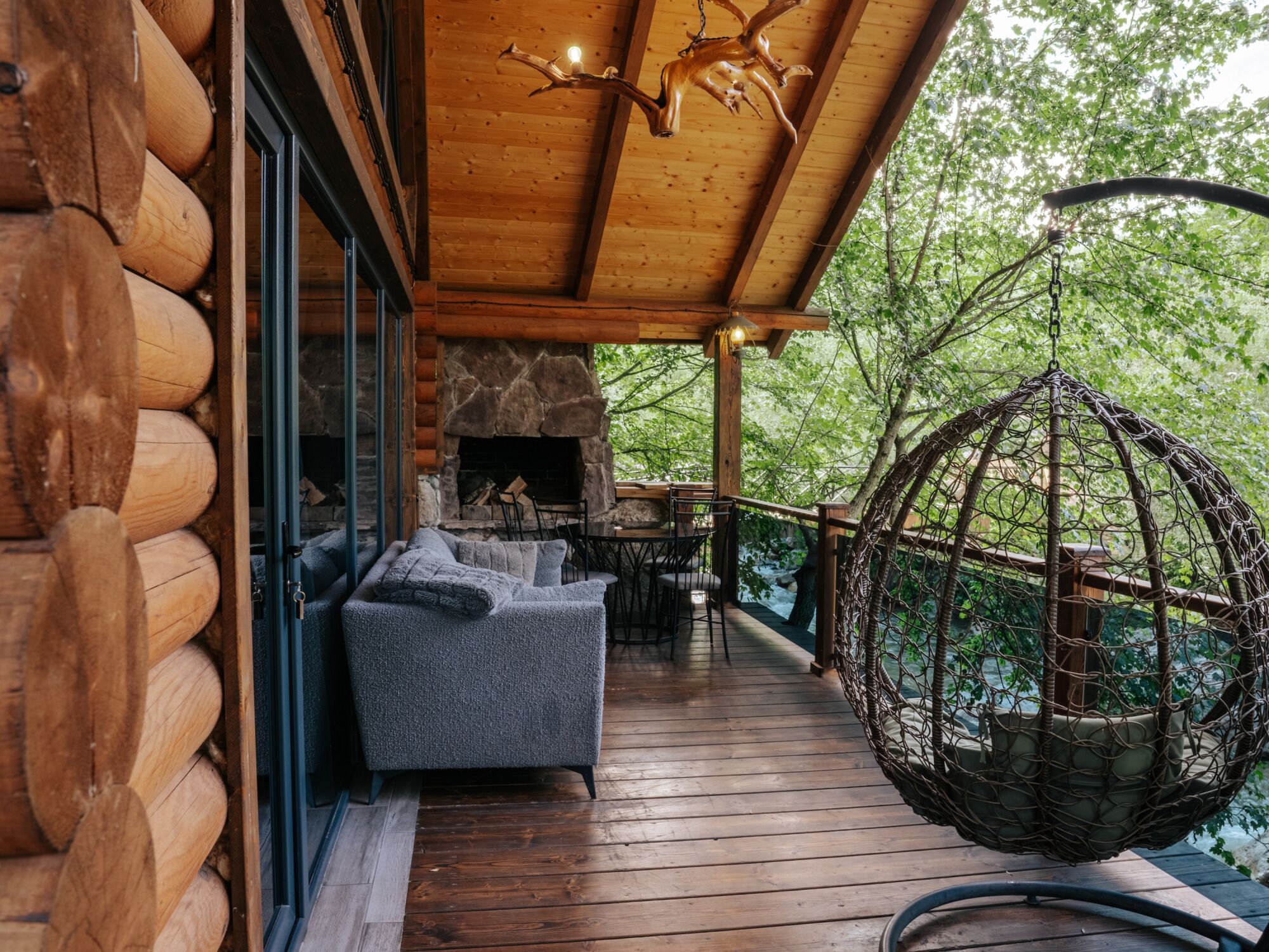 A wooden deck with a stone fireplace, outdoor seating, and a hanging chair overlooking a forested area.