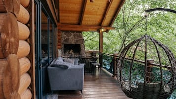 A wooden deck with a stone fireplace, outdoor seating, and a hanging chair overlooking a forested area.