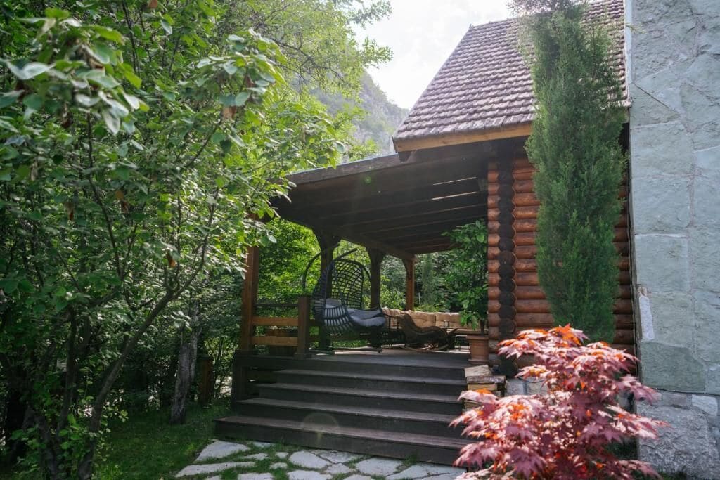 A wooden gazebo with a stone wall, surrounded by lush greenery and a stone pathway.