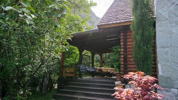 A wooden gazebo with a stone wall, surrounded by lush greenery and a stone pathway.