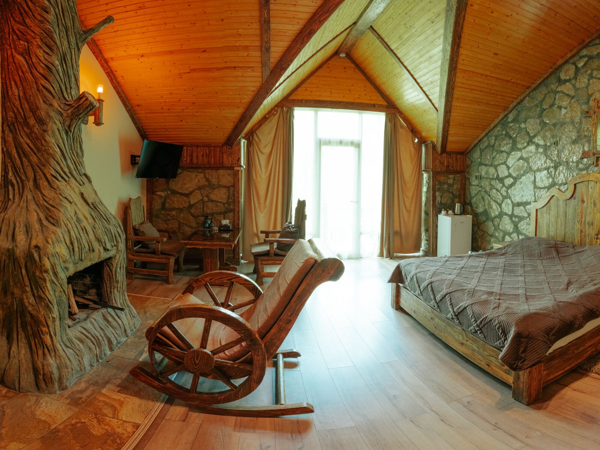 Une chambre à coucher rustique avec une cheminée en pierre, un fauteuil à bascule en bois et un lit recouvert d’une courtepointe.