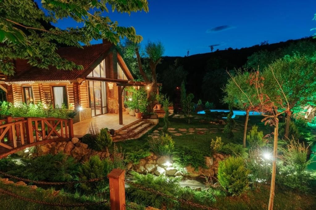 A wooden cabin with a porch, surrounded by a landscaped garden with a small stream and a bridge.