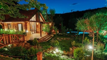 A wooden cabin with a porch, surrounded by a landscaped garden with a small stream and a bridge.