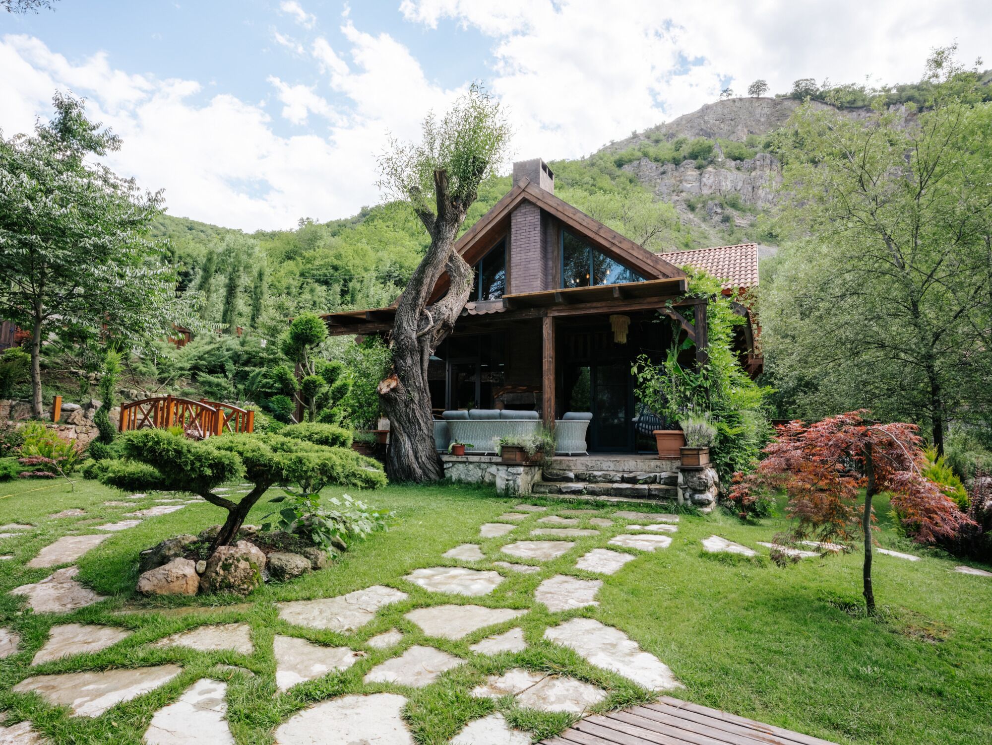 A rustic mountain cabin with a stone pathway leading to the entrance, surrounded by lush greenery and a view of the mountain.