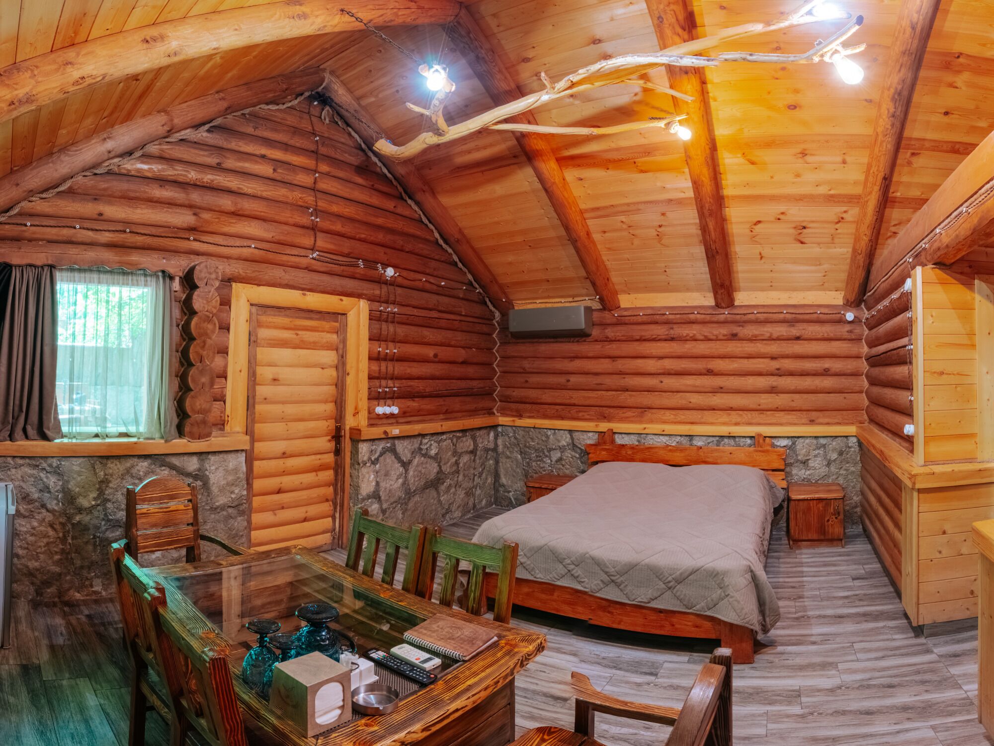 A log cabin interior with a bed, a table with chairs, a window with curtains, and a stone wall.
