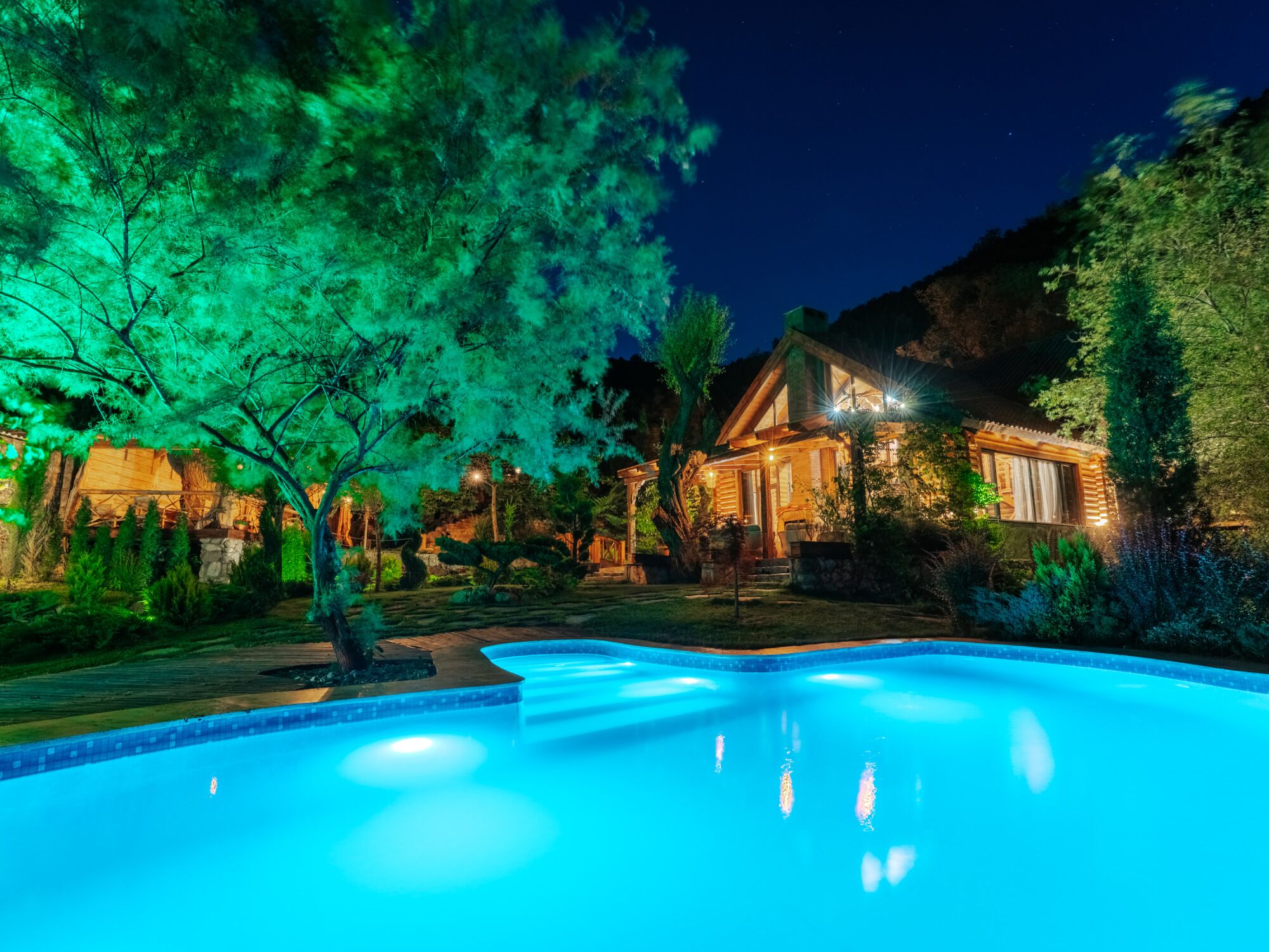 A swimming pool with a wooden deck, surrounded by trees and a house with illuminated windows.