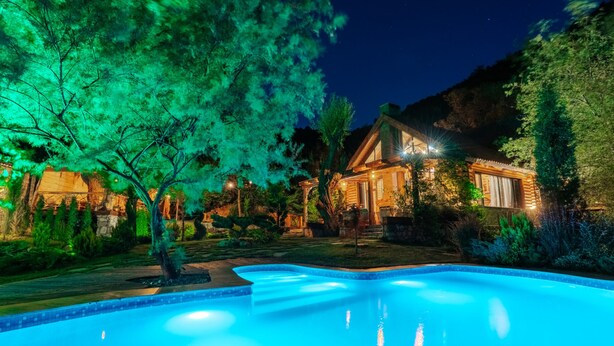 A swimming pool with a wooden deck, surrounded by trees and a house with illuminated windows.