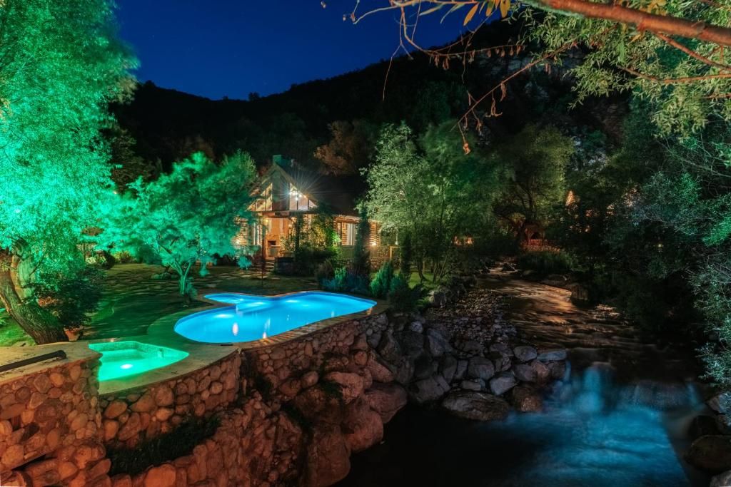 A stone pool with a waterfall, surrounded by trees and a lit-up house in the background.