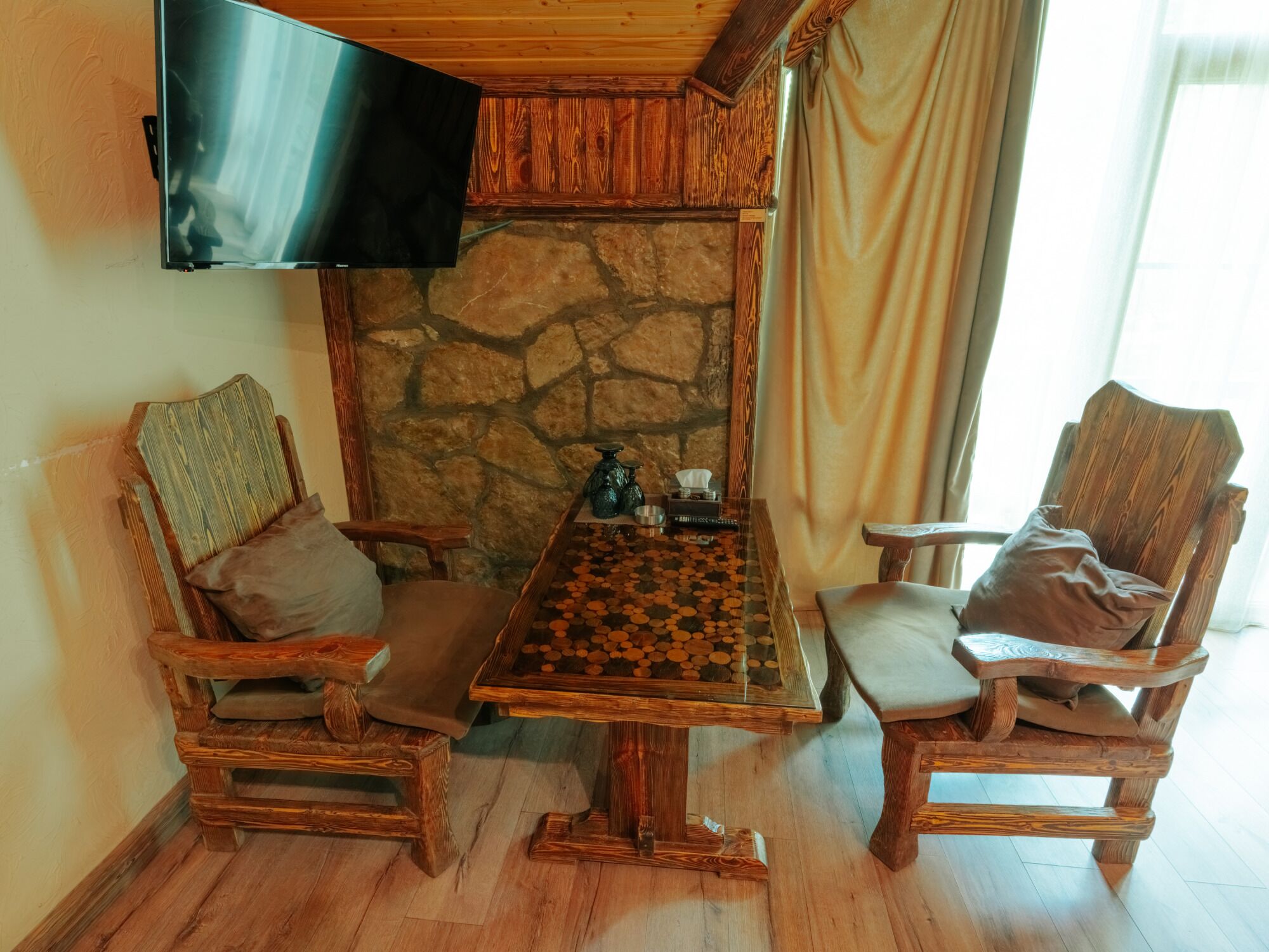 A wooden dining table with a chessboard, flanked by two wooden chairs with cushions. A stone wall and a television mounted on the wall are also visible.