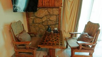 A wooden dining table with a chessboard, flanked by two wooden chairs with cushions. A stone wall and a television mounted on the wall are also visible.