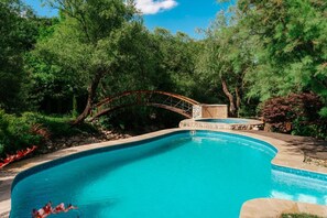 A swimming pool with a stone border, surrounded by lush greenery and a wooden bridge.