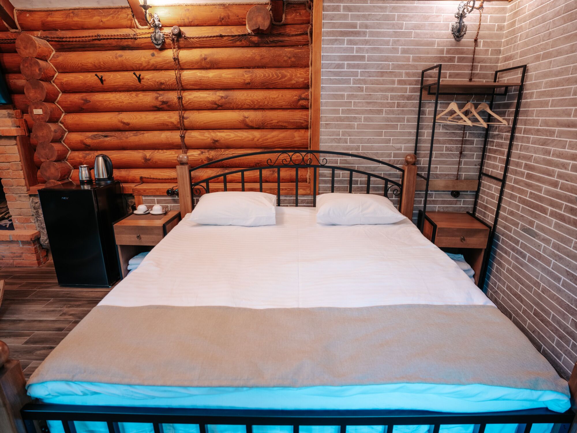 Una cama doble con cabecera de madera, una mesita de noche con tetera y un perchero.