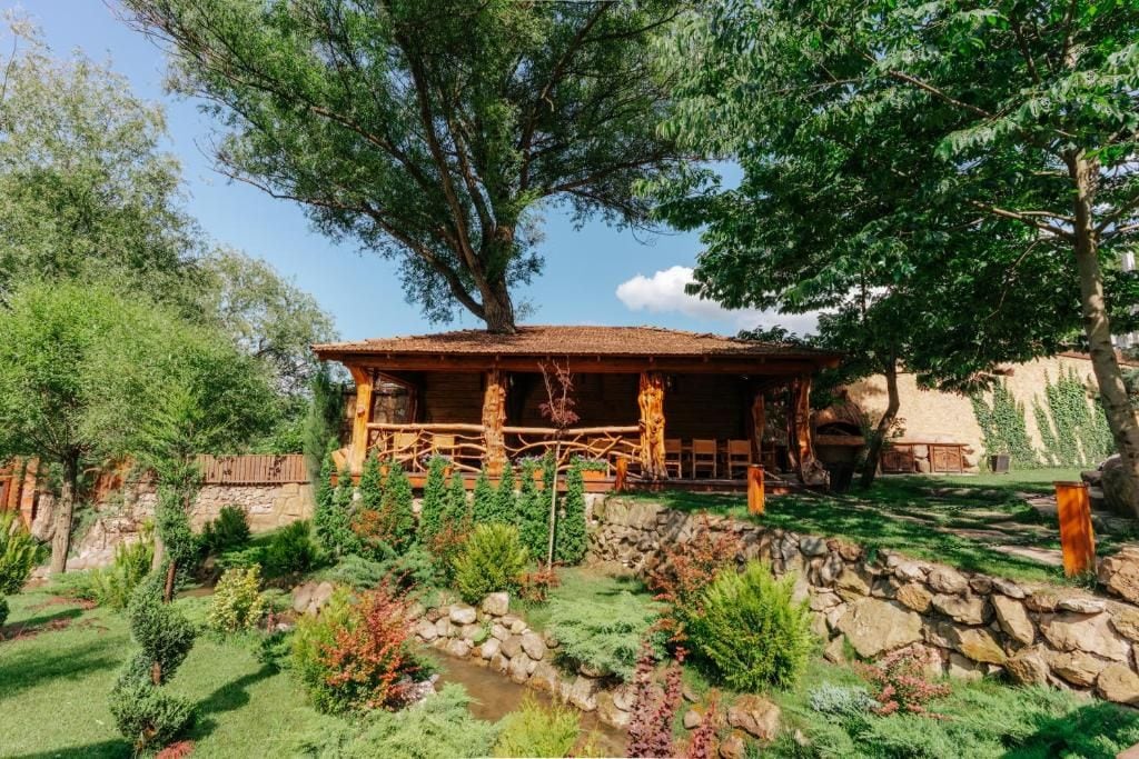 Una cabaña de madera rústica con techo de paja, rodeada por un jardín con diversas plantas y un pequeño arroyo.