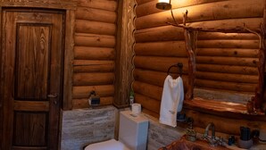 A wooden cabin bathroom with a toilet, sink, and a wooden shelf with toiletries.