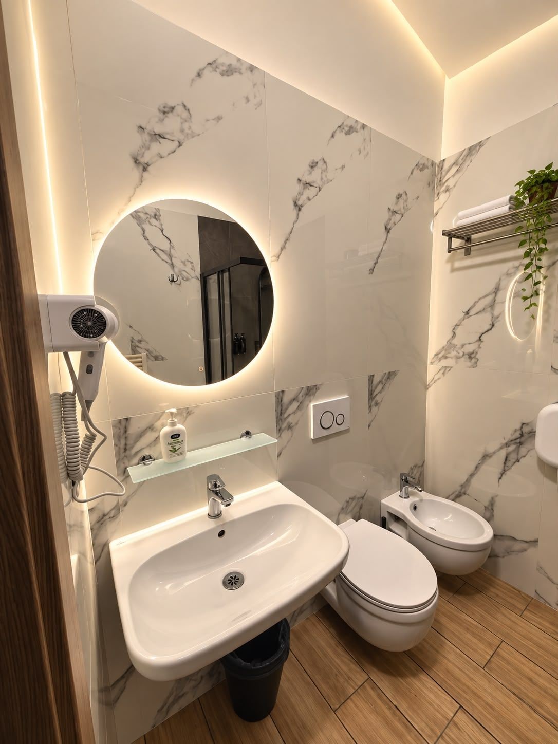 Une salle de bain moderne avec un pare-feu en marbre, un grand miroir rond doté d’un éclairage intégré, un sèche-cheveux, un lavabo, un bidet et une petite étagère contenant des articles de toilette.