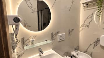 Une salle de bain moderne avec un pare-feu en marbre, un grand miroir rond doté d’un éclairage intégré, un sèche-cheveux, un lavabo, un bidet et une petite étagère contenant des articles de toilette.