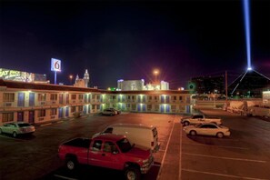 A hotel with multiple floors, a parking lot with several cars, and a prominent building with a lit sign in the background.
