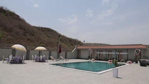 Zona de piscina exterior con sillones de playa y sombrillas, un edificio con un cartel, y un paisaje montañoso al fondo.