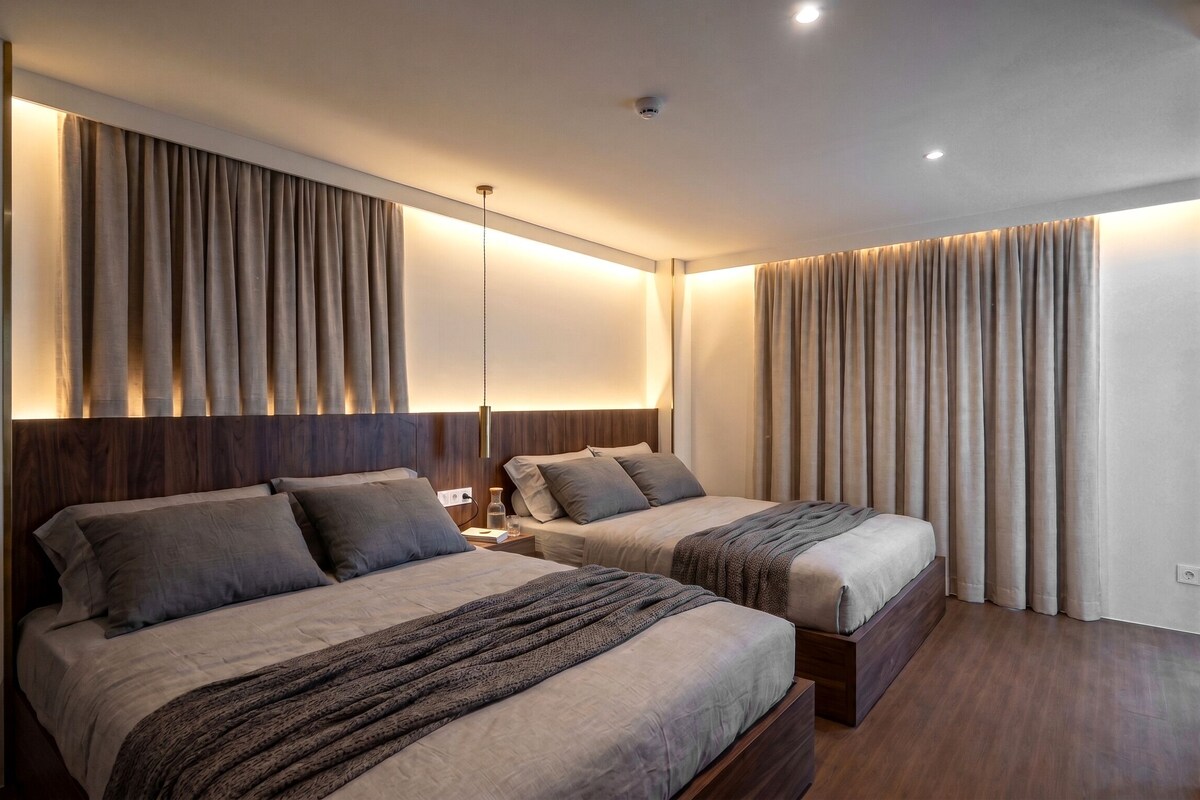 Una camera d'albergo con due letti, una testiera in legno, un comodino con una lampada e un'ampia finestra con tende.